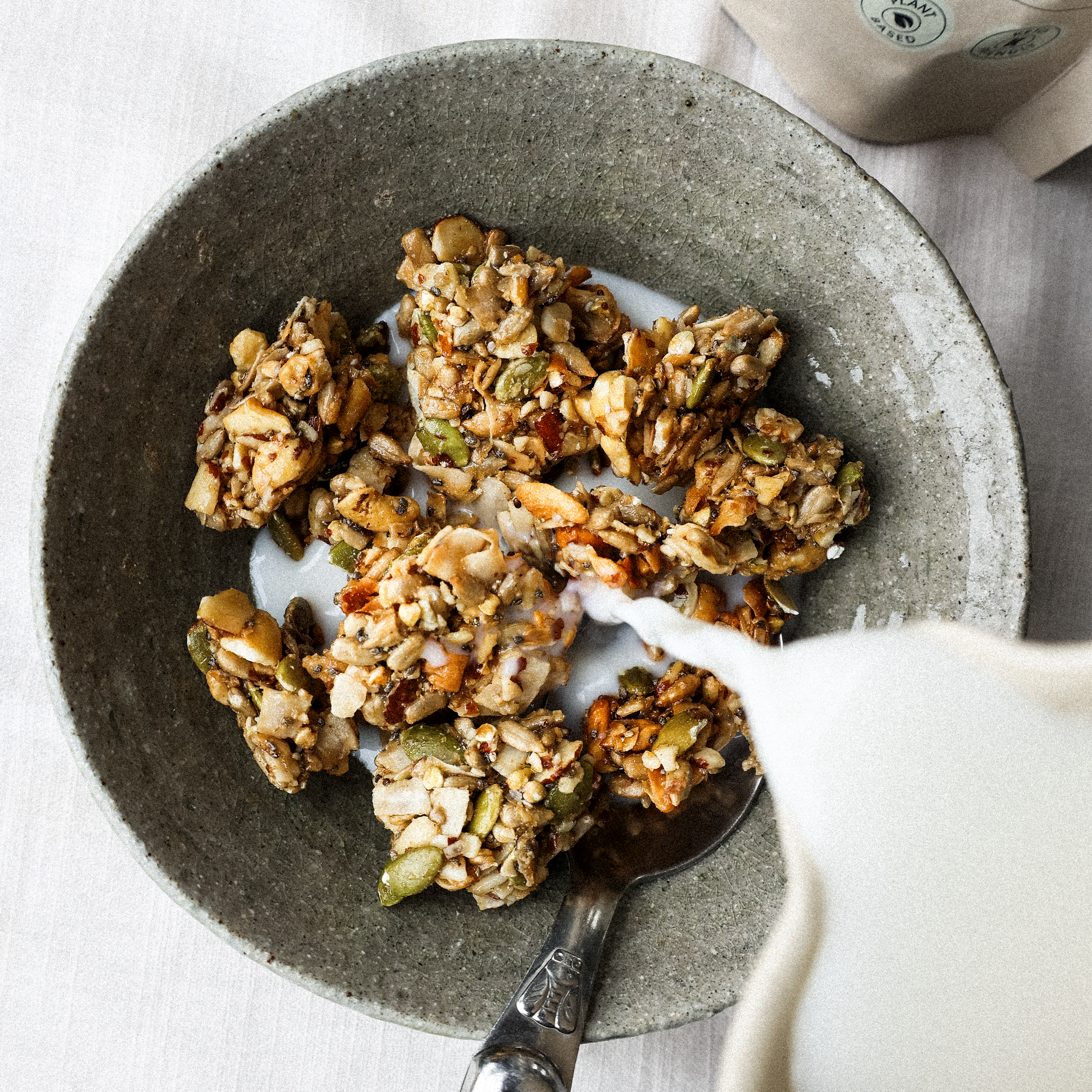 Milk pouring over Hapüs Grain-Free Granola and a spoon in a textured ceramic bowl.